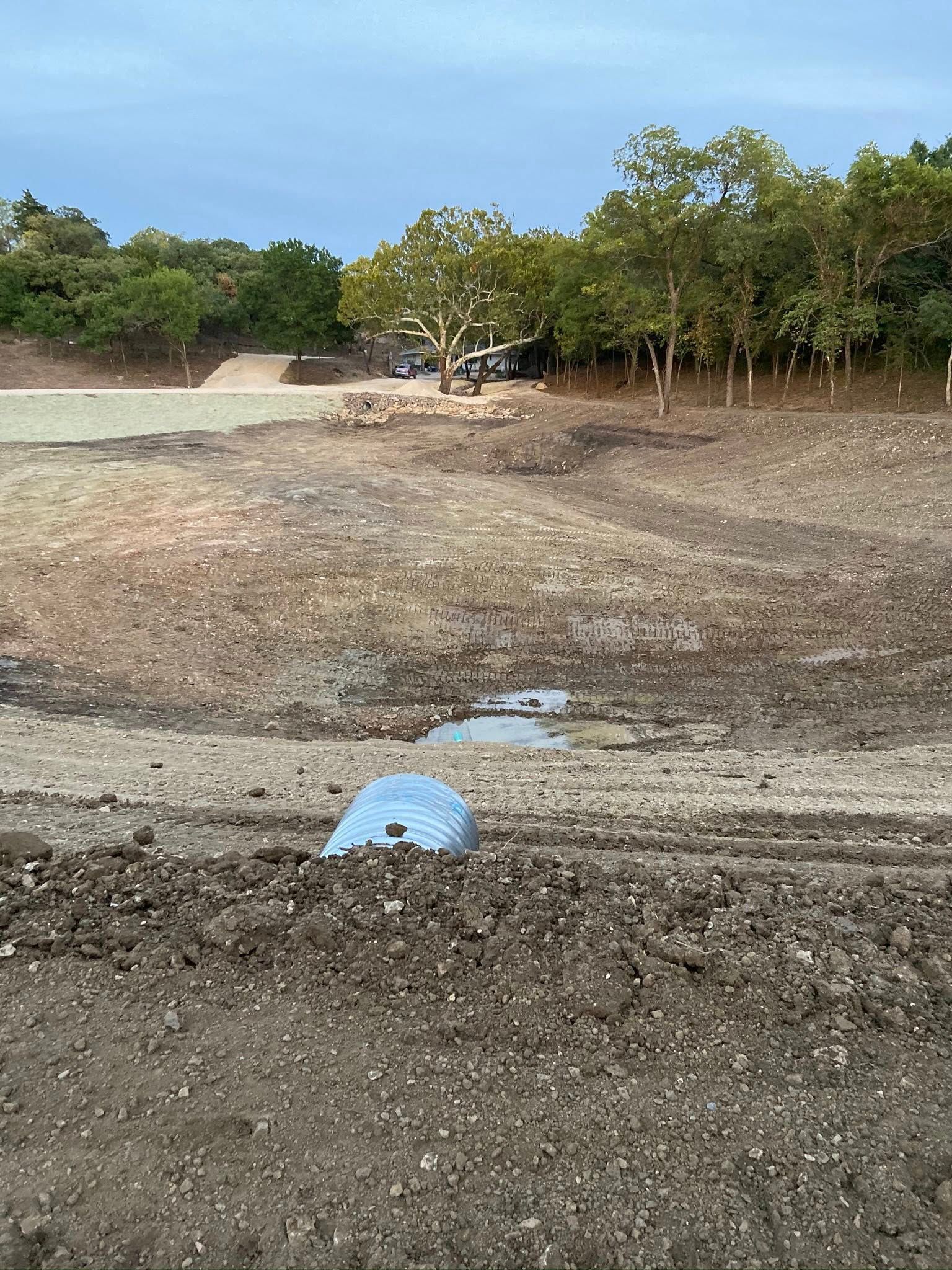 Dry dirt field with a blue pipe in the foreground and trees along the edge under a cloudy sky