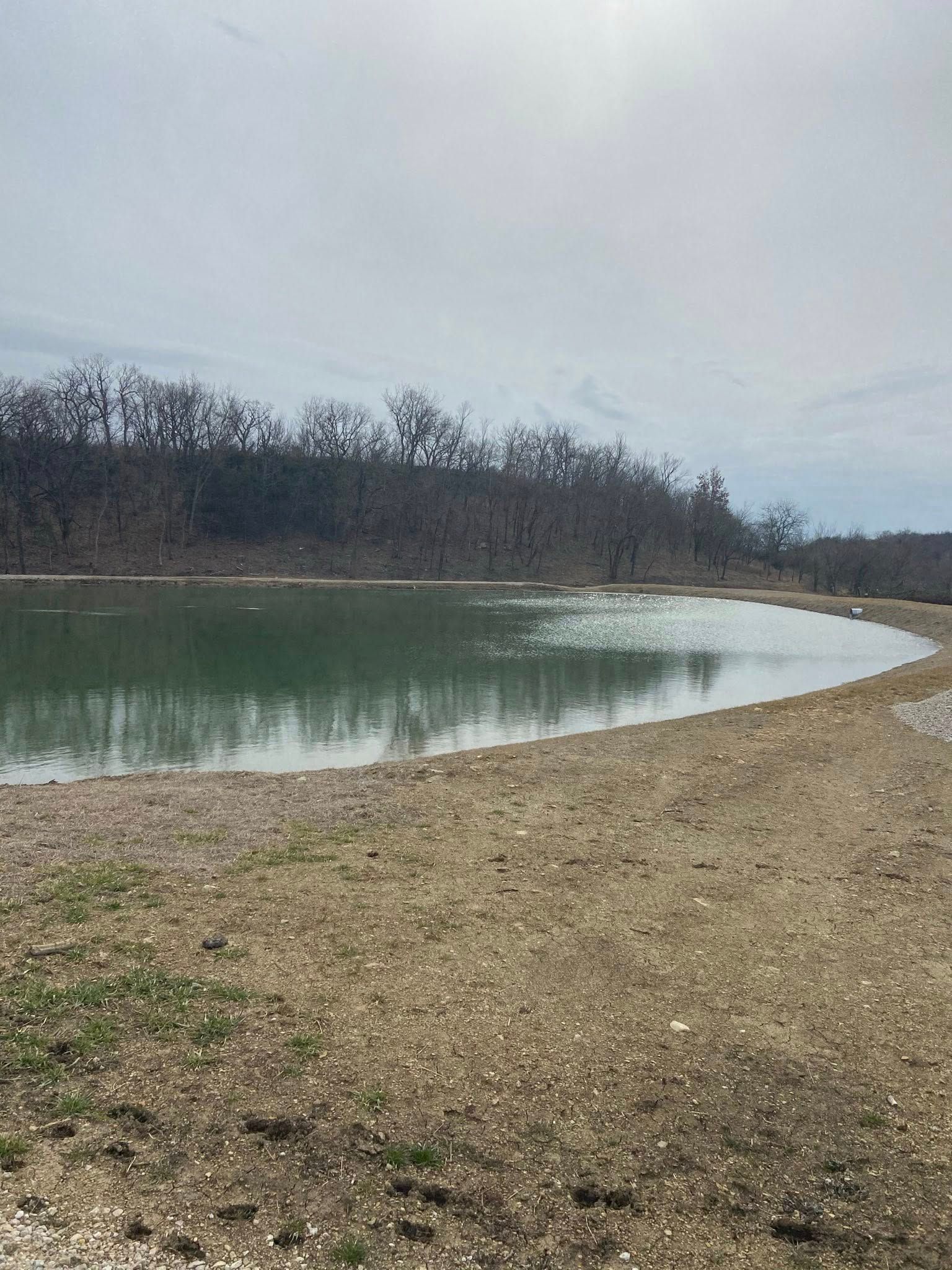 Calm river beside a gravel bank under a cloudy sky, with leafless trees along the far shore