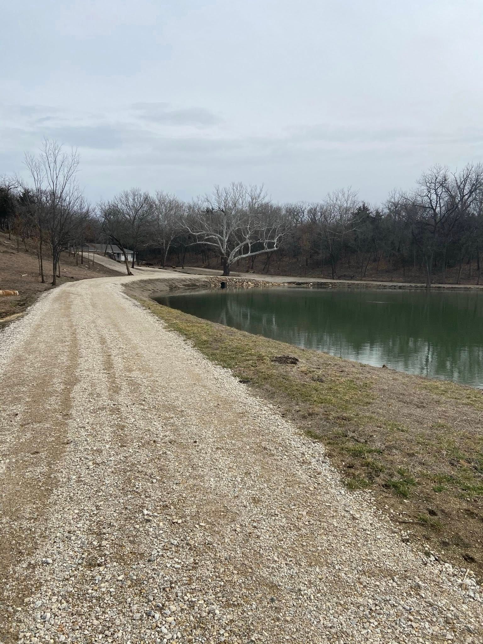 Gravel path beside a calm green pond under a cloudy sky, with bare trees in the distance