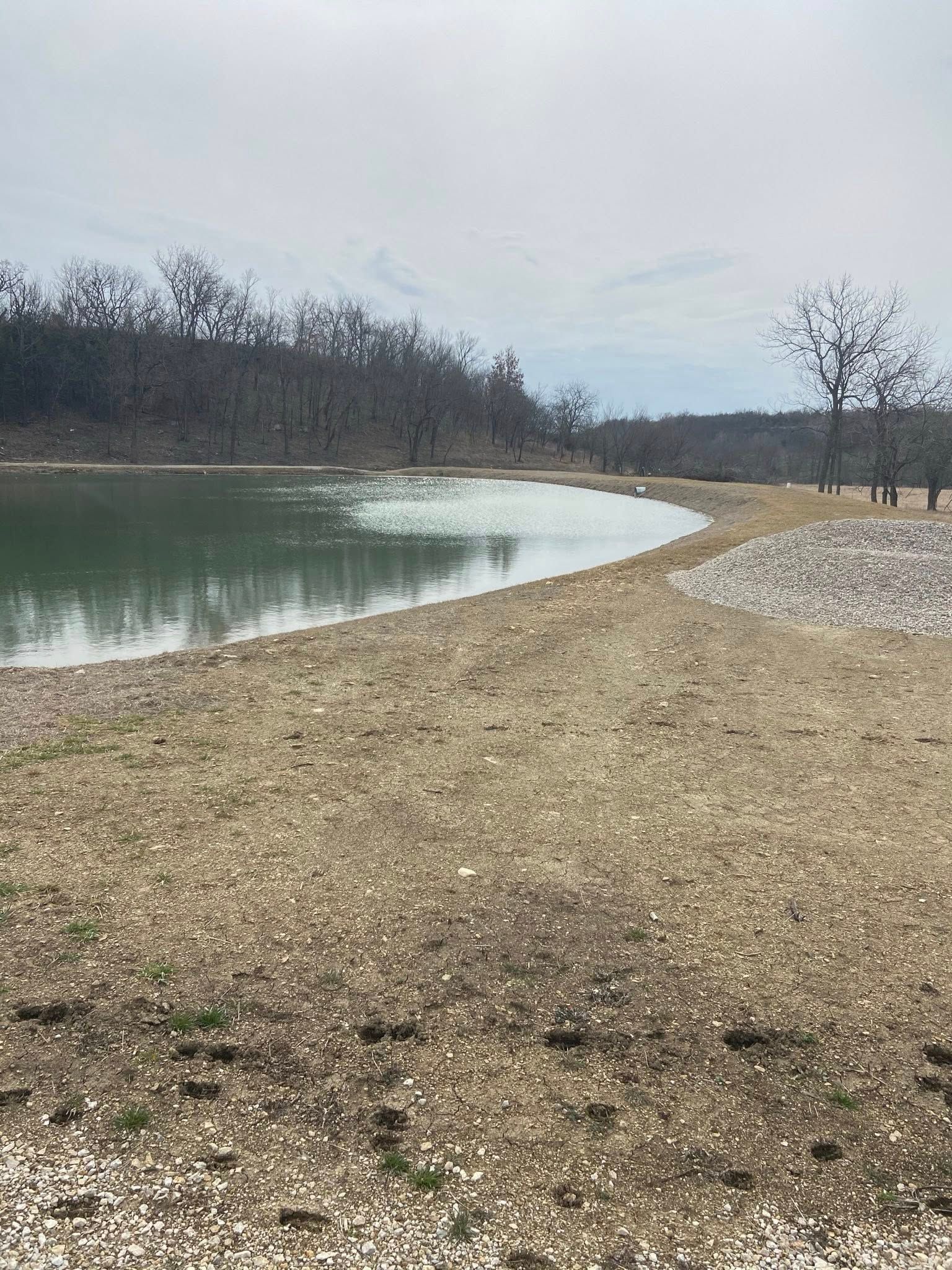 Curved gravel shoreline beside a calm lake, with bare trees and cloudy sky in the background