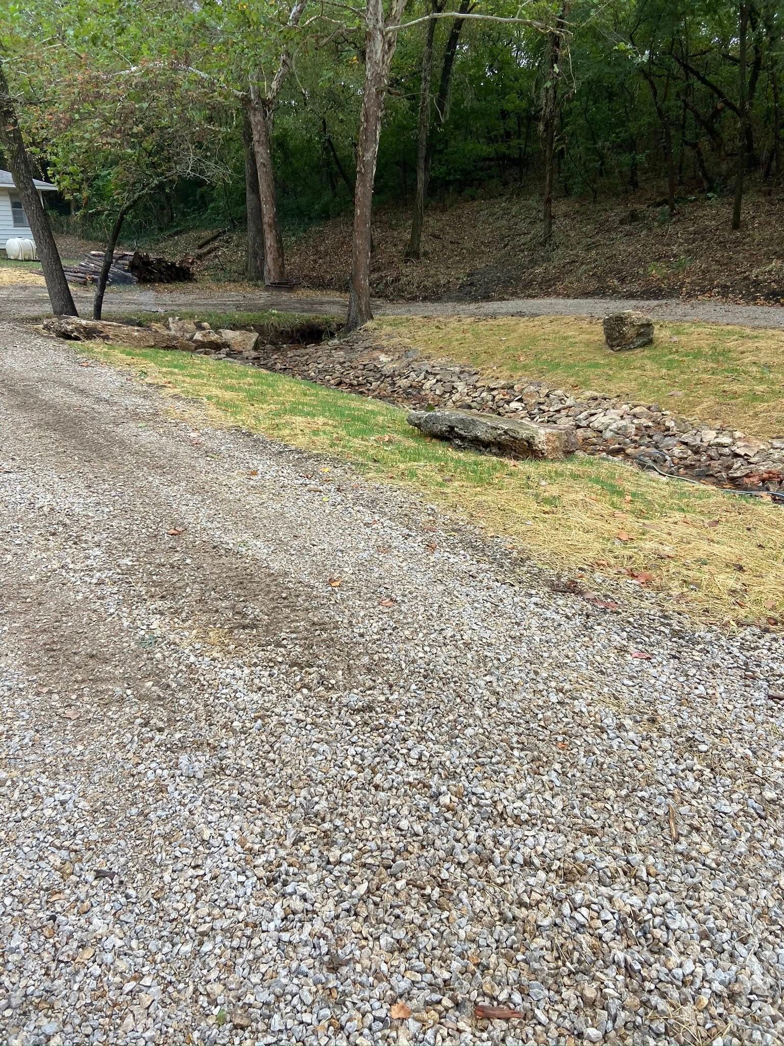 Gravel driveway beside a stone border and trees in a wooded yard