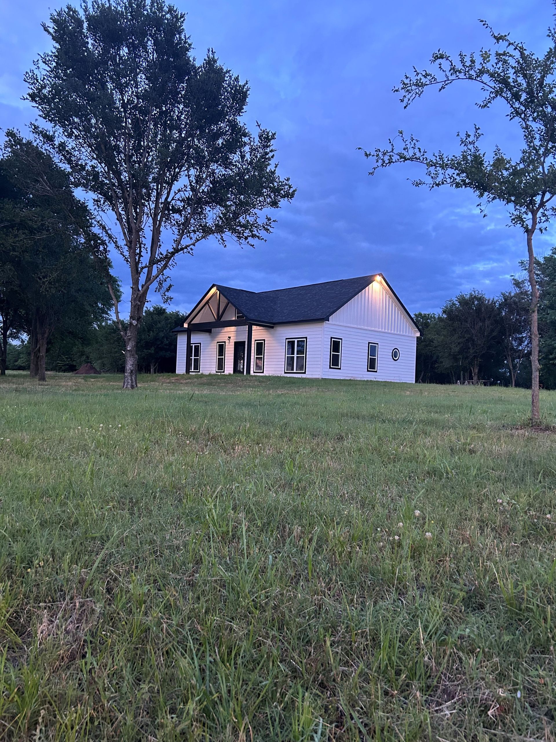 Small white house with lit windows at dusk, surrounded by a grassy field and tall trees.