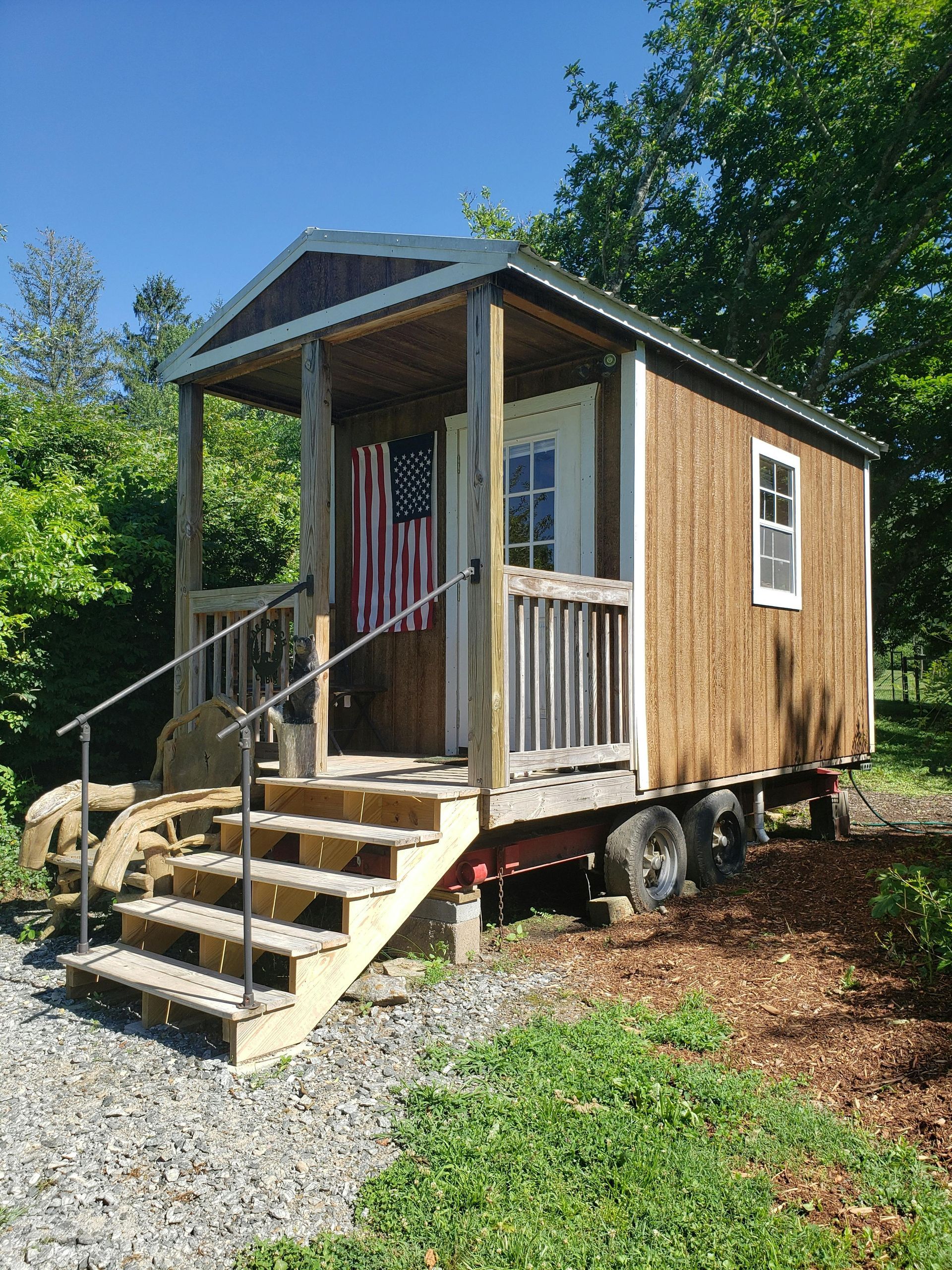 Small wooden cabin on wheels with front steps and porch, set on a sunny gravel lot with trees nearby