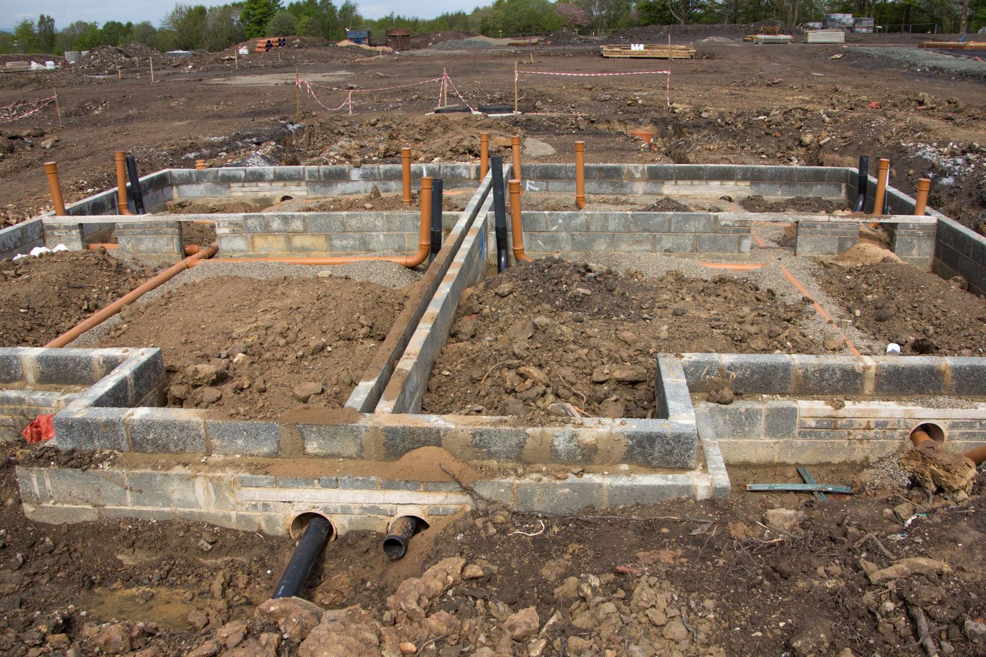 Foundation trenches and concrete footings at a construction site with exposed pipes and rebar.