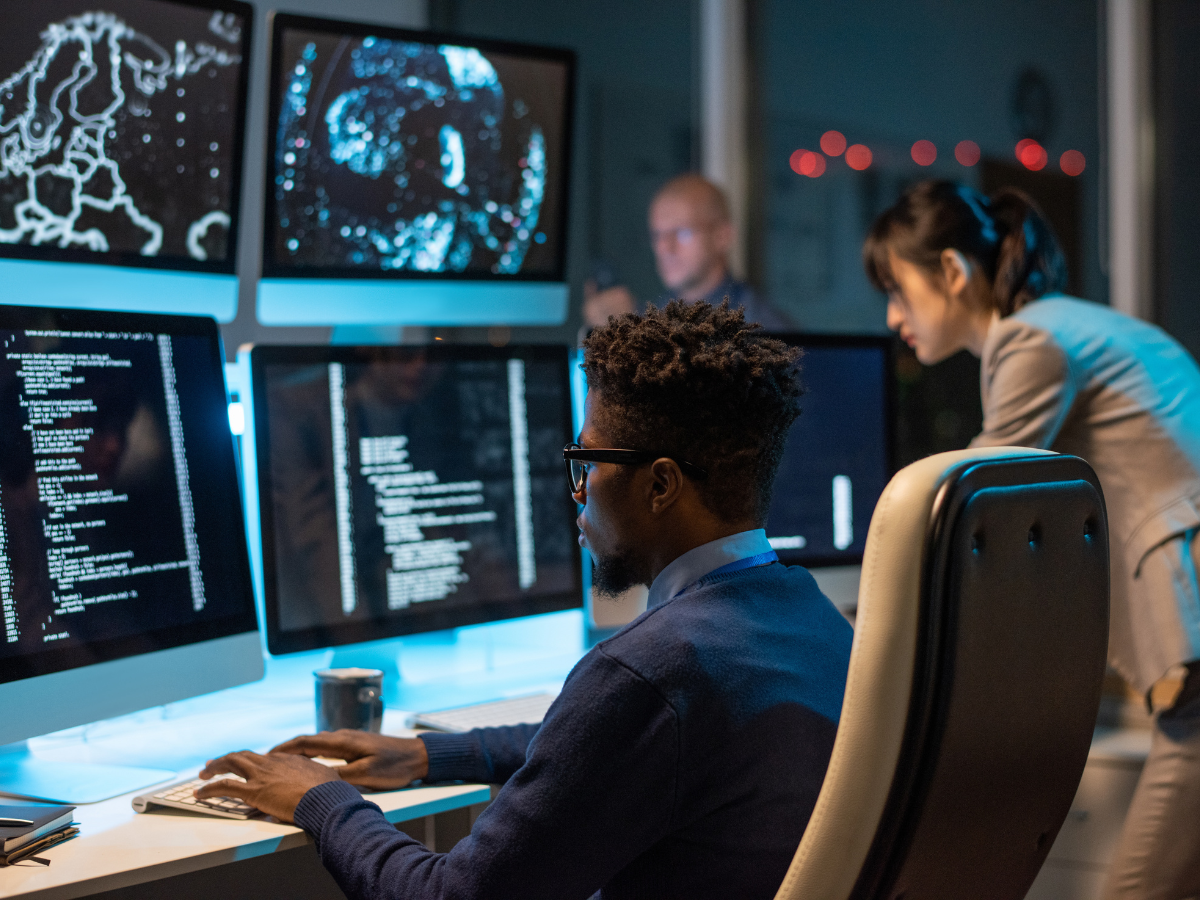 Man coding at a computer, other team members in a dark office with multiple monitors displaying code and data.