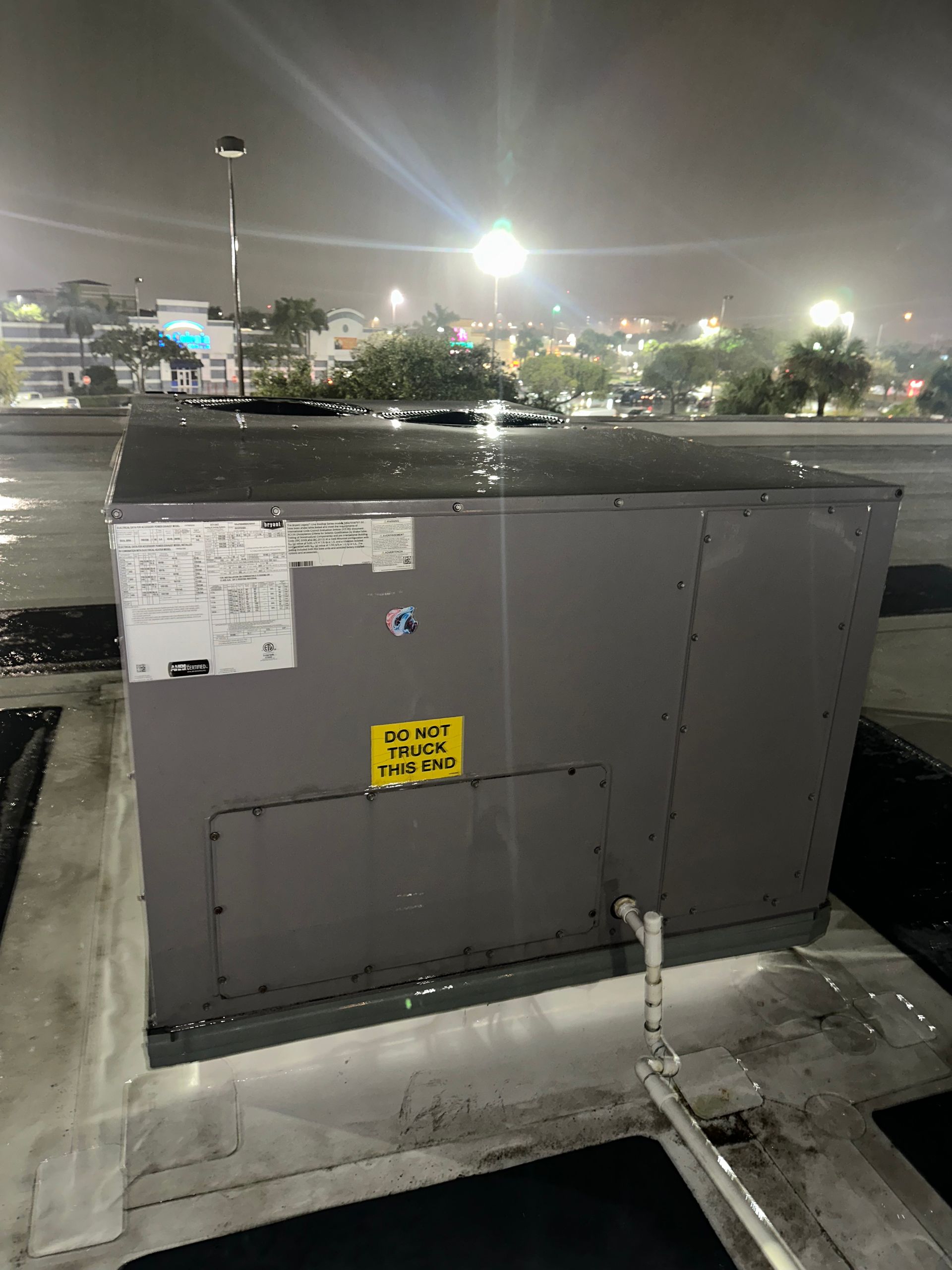 Gray utility box on a wet parking lot curb at night with a warning label and nearby trees in the background