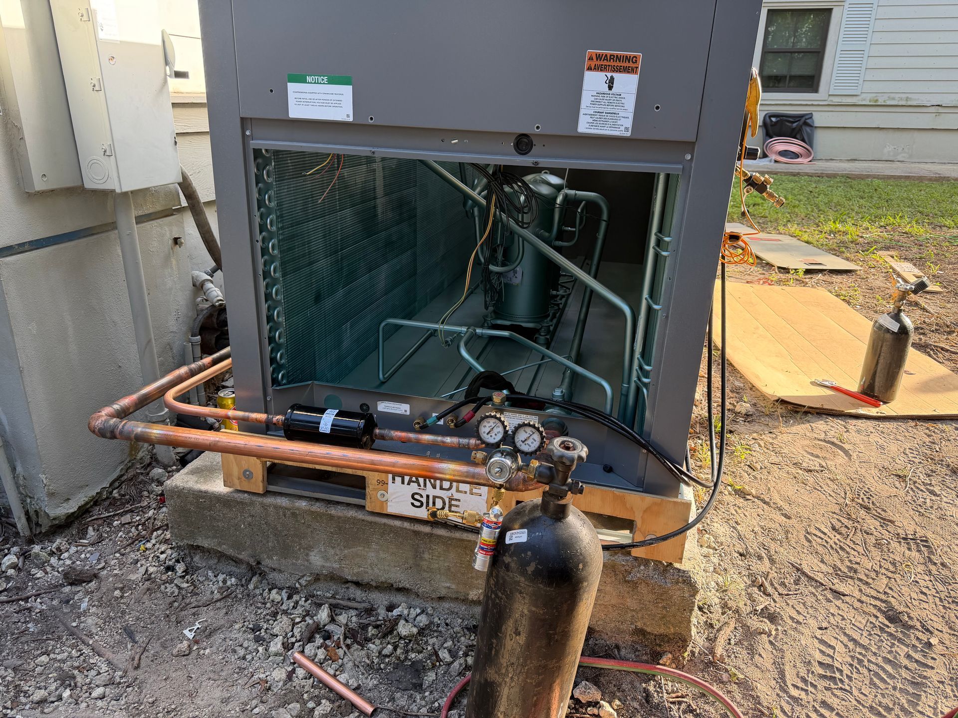 Outdoor HVAC unit with exposed copper pipes and a black refrigerant cylinder attached in front