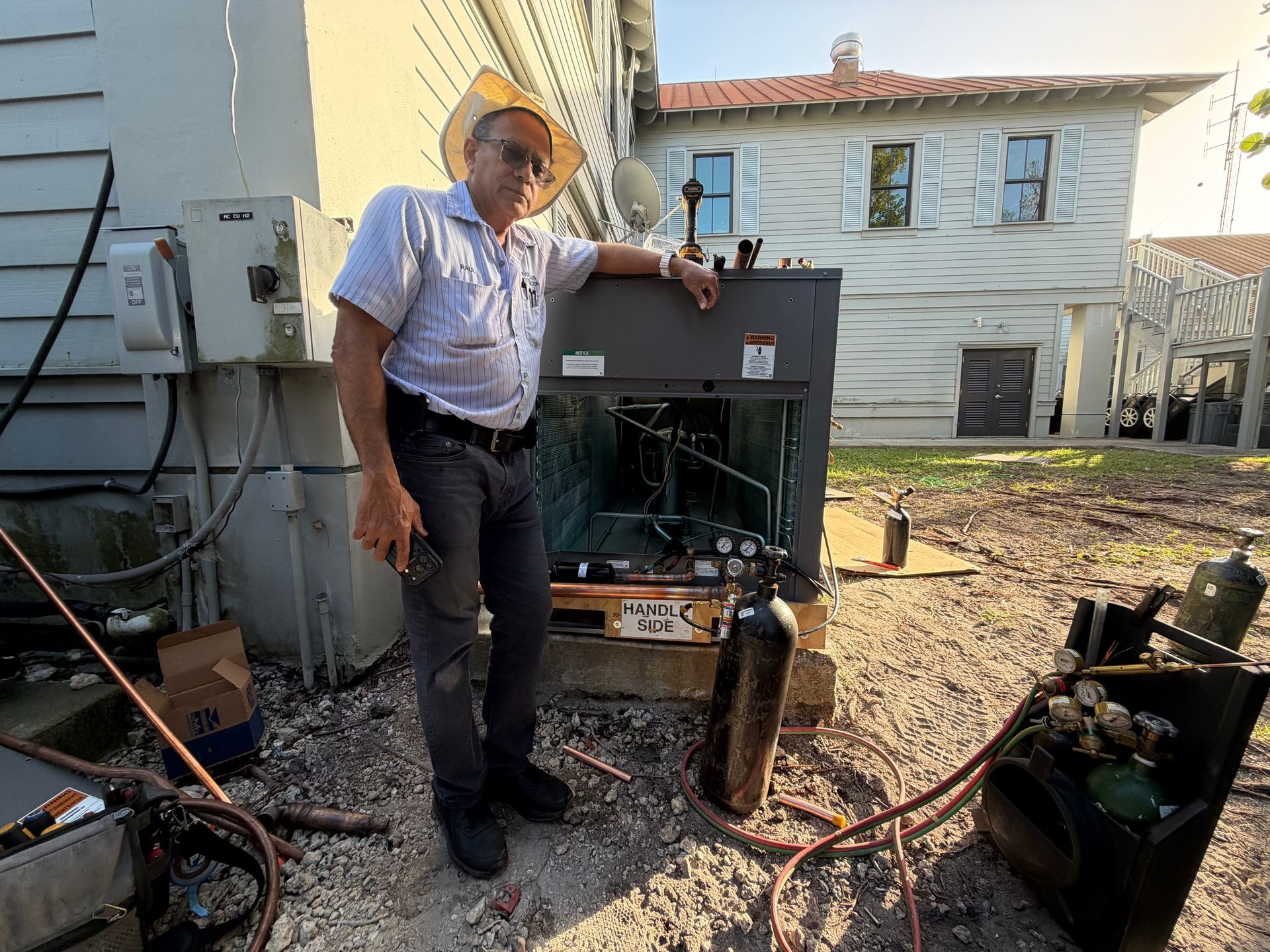 Man standing beside an open generator outdoors, with tools and cables on the ground рядом a building