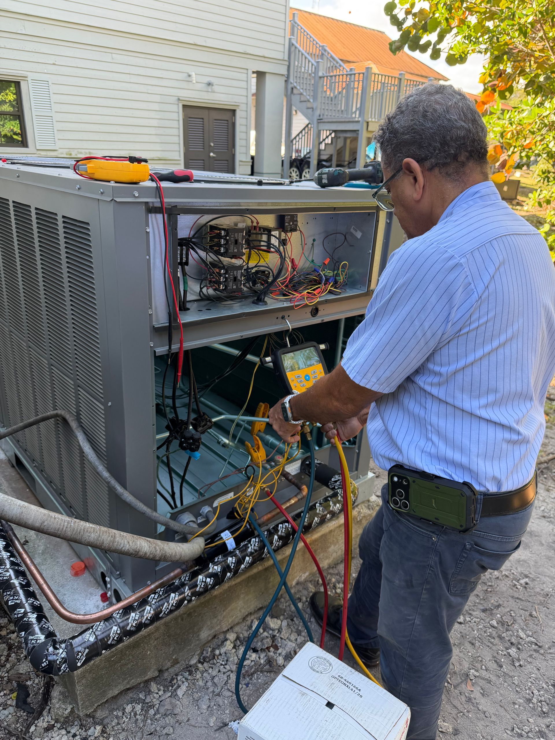 Technician working on exposed HVAC unit outdoors, holding gauges and wires near the open panel