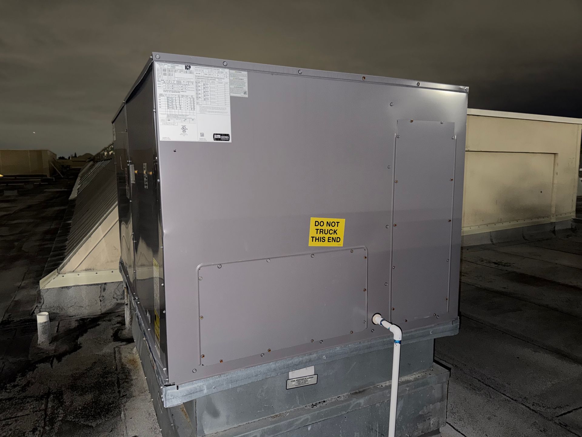 Large gray rooftop HVAC unit with warning label and white pipe at night