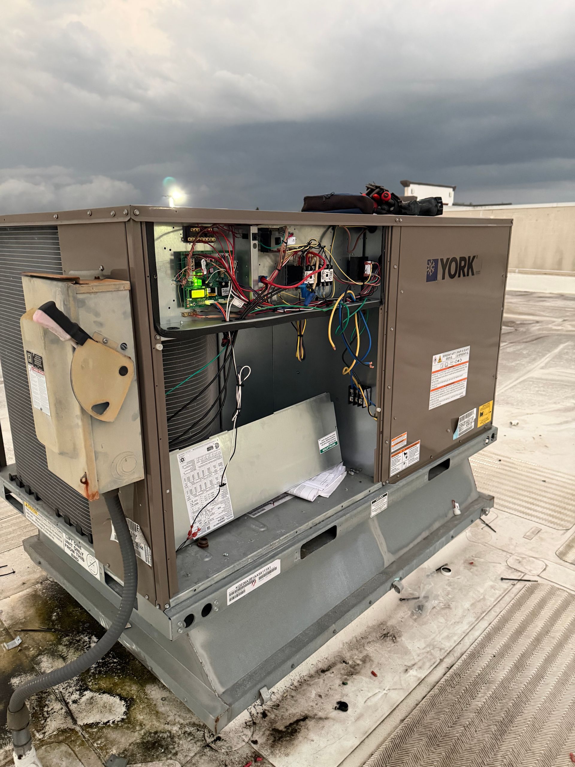 Open electrical HVAC unit with exposed wiring on a rooftop under cloudy sky