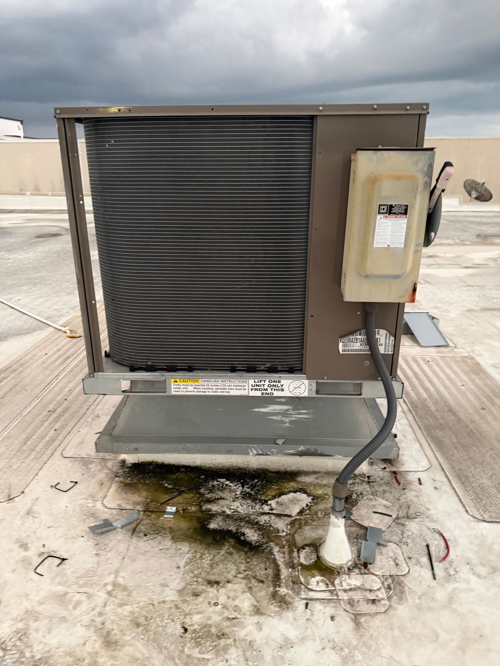 Outdoor HVAC unit with exposed coils and a beige control box on a concrete rooftop.