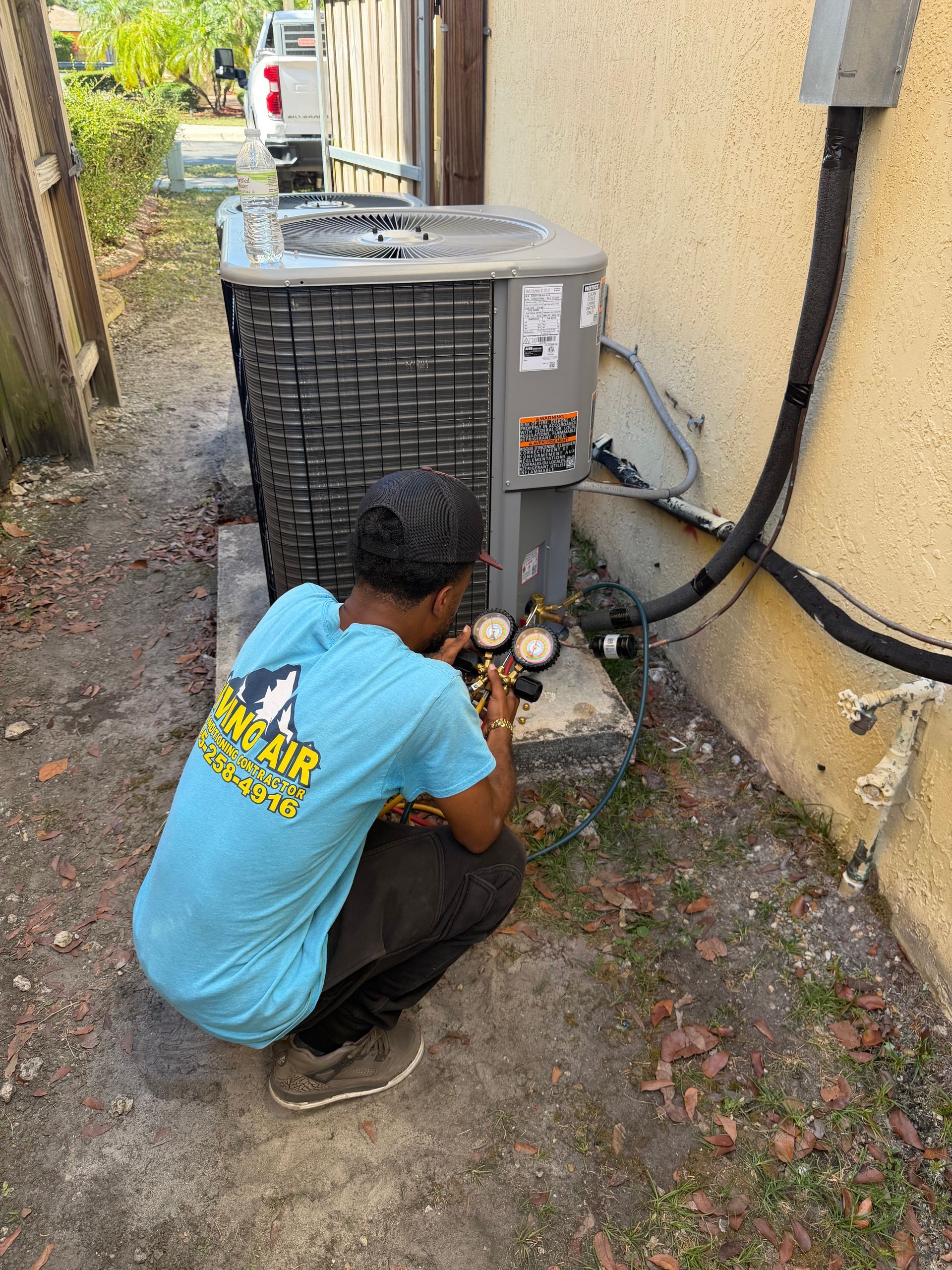 Technician crouching beside an outdoor HVAC unit, checking gauges in a narrow alley.