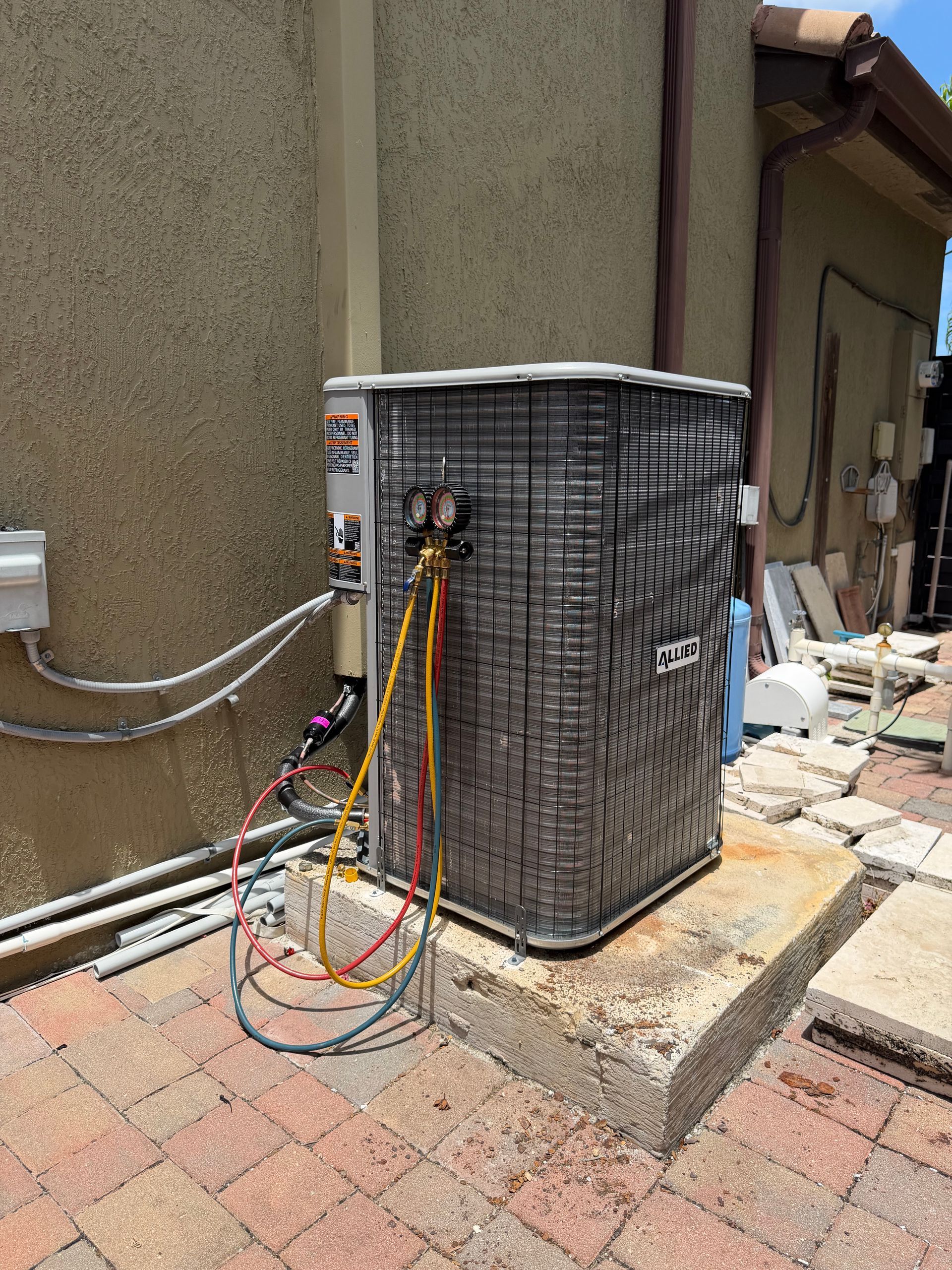 Outdoor HVAC unit with exposed colorful wiring beside a stucco house wall and brick patio