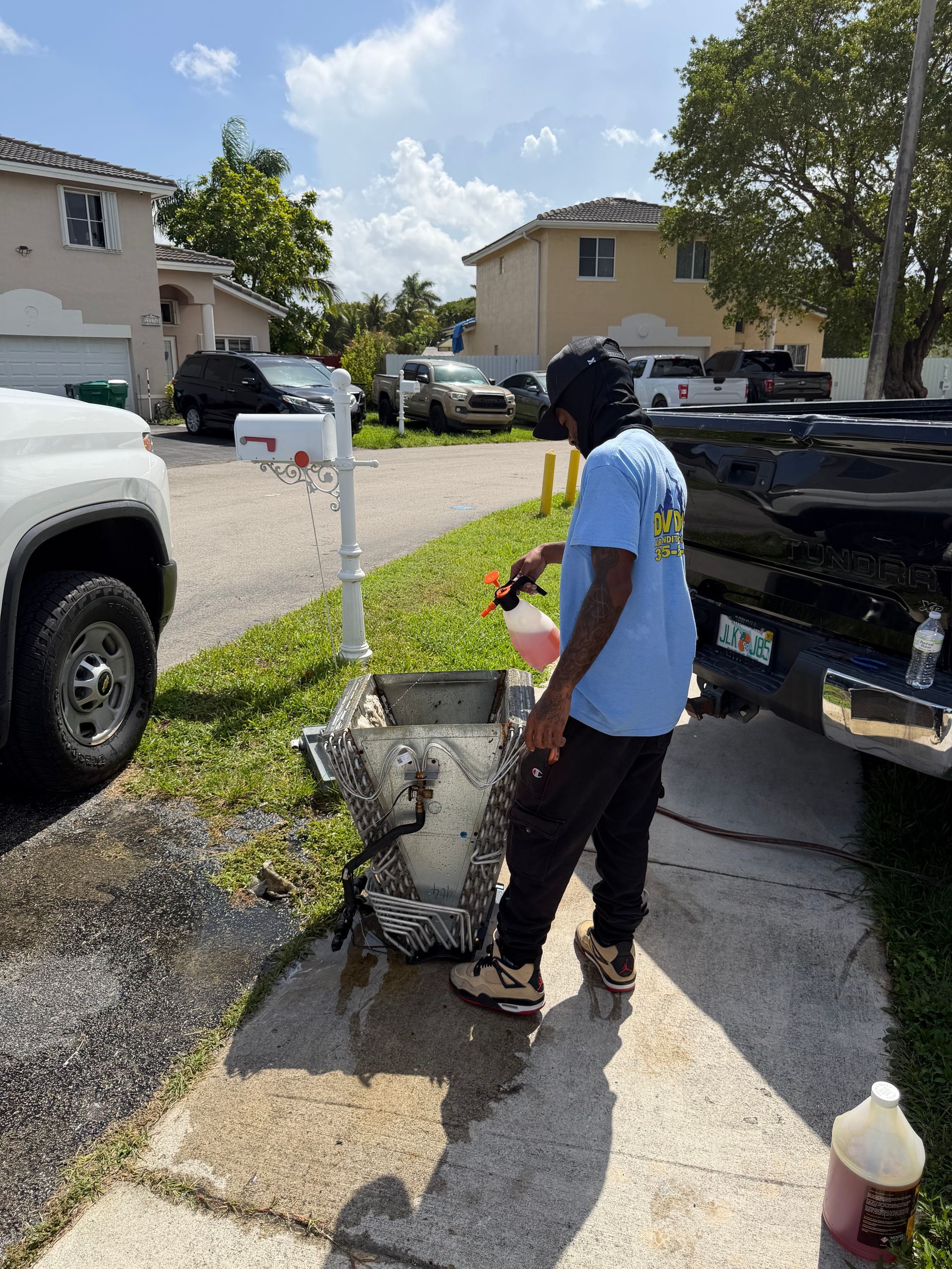 Person washing a black pickup truck on a driveway with a hose and bucket in a sunny neighborhood.