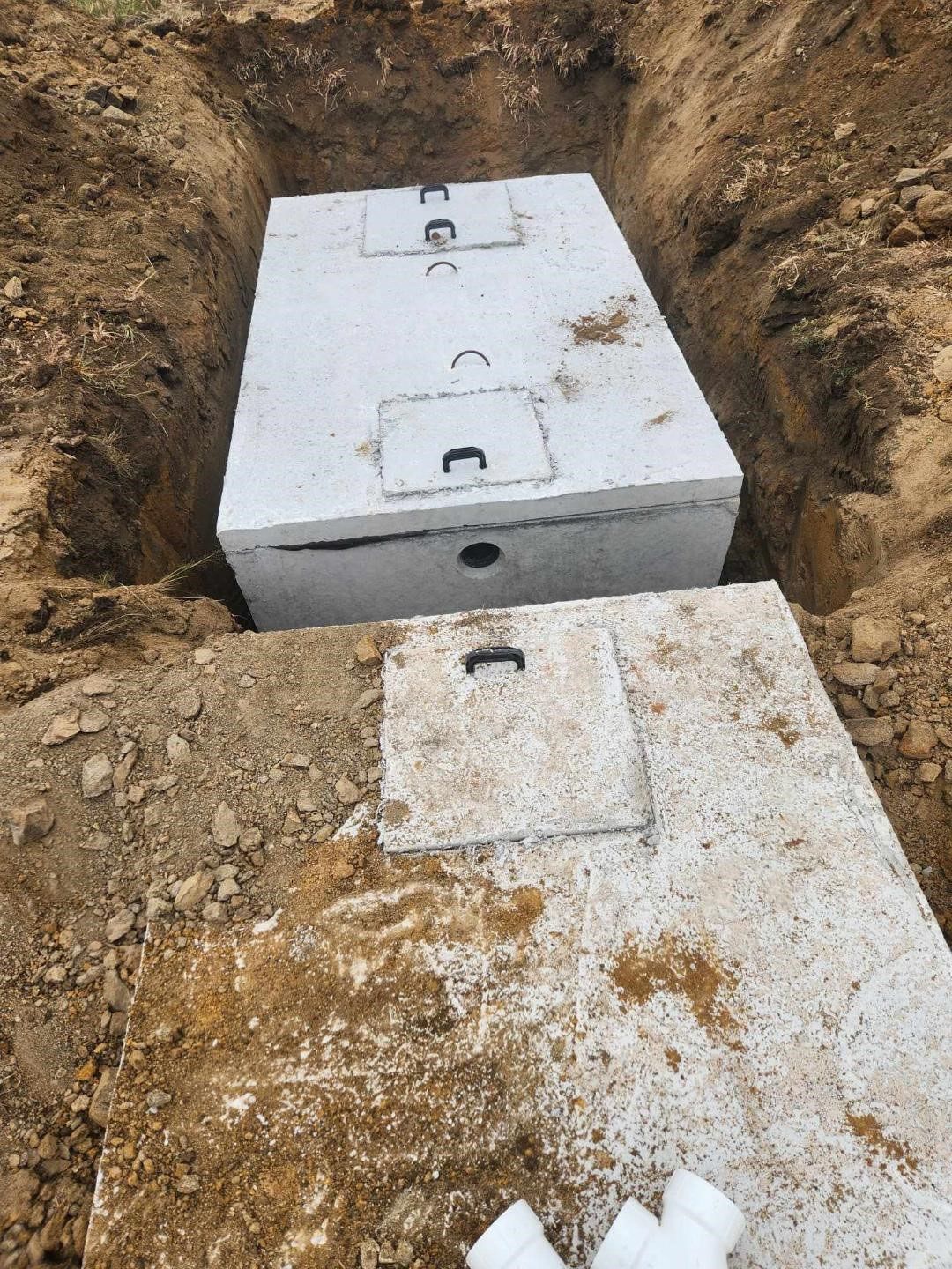 Concrete septic tank in an excavated trench, lid removed, and another lid lying nearby.