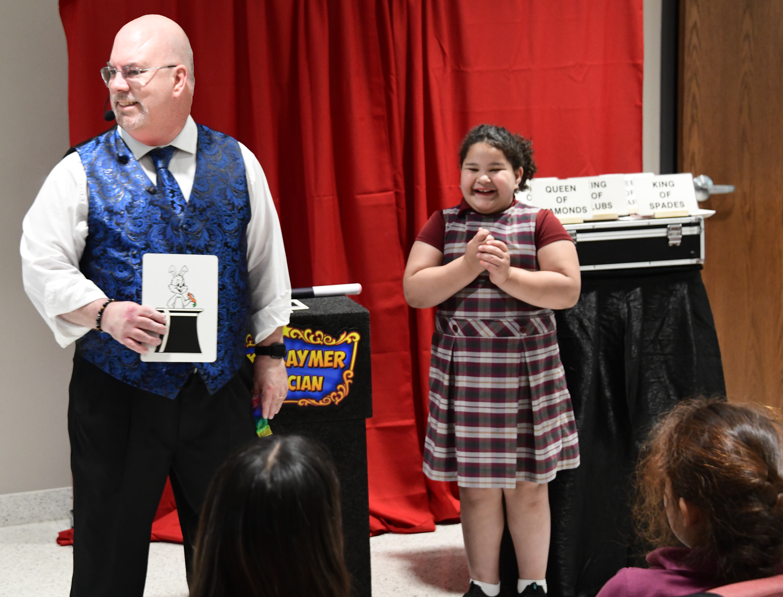 Magician and young girl on stage, magician holding a picture of a rabbit, audience watches.