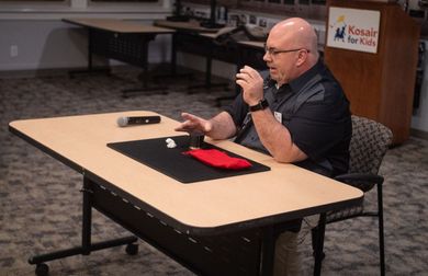 Man seated at table, gesturing; small objects and red cloth visible. 