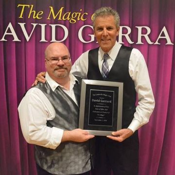 Two men with a plaque on a stage with a banner that reads 