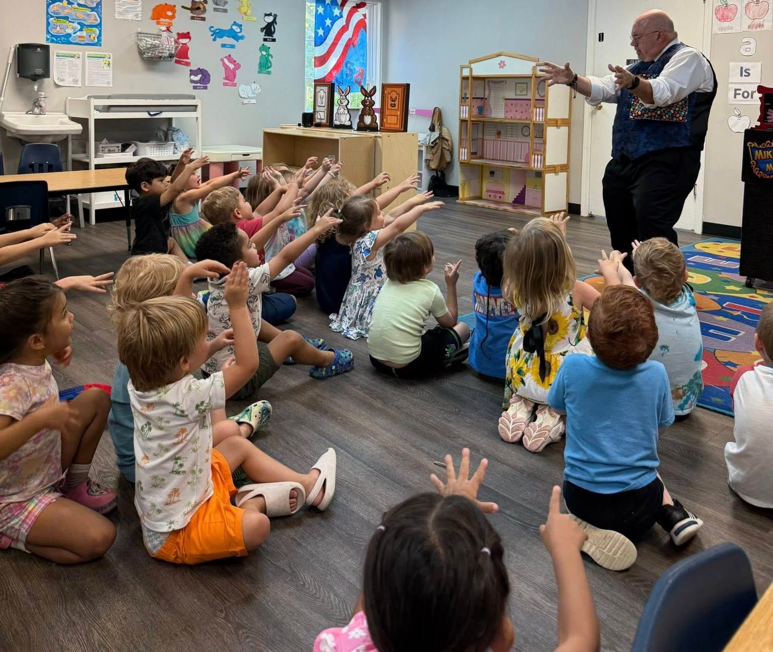 Children in a circle watch a magician perform in a classroom. They have their hands up in the air.
