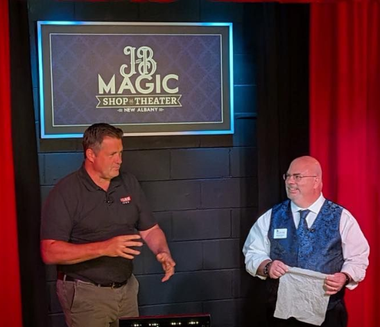 Two men on stage, one gesturing, the other holding a cloth. Stage backdrop: 