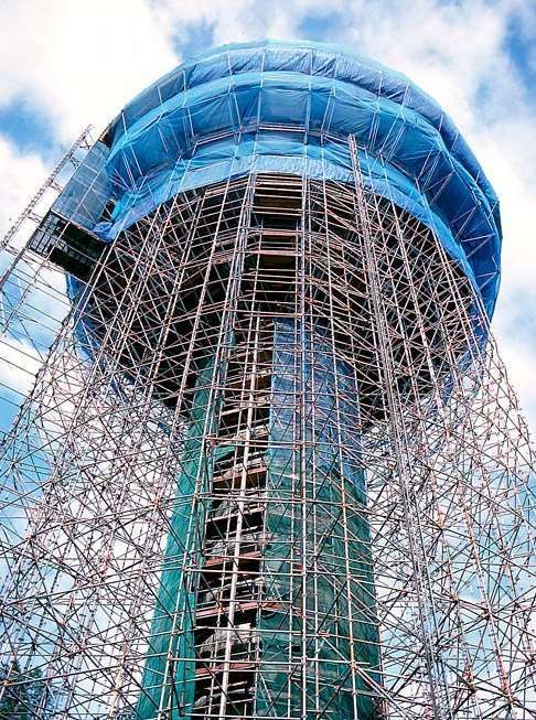 Torre de aço em construção com andaimes azuis e topo arredondado contra um céu nublado.