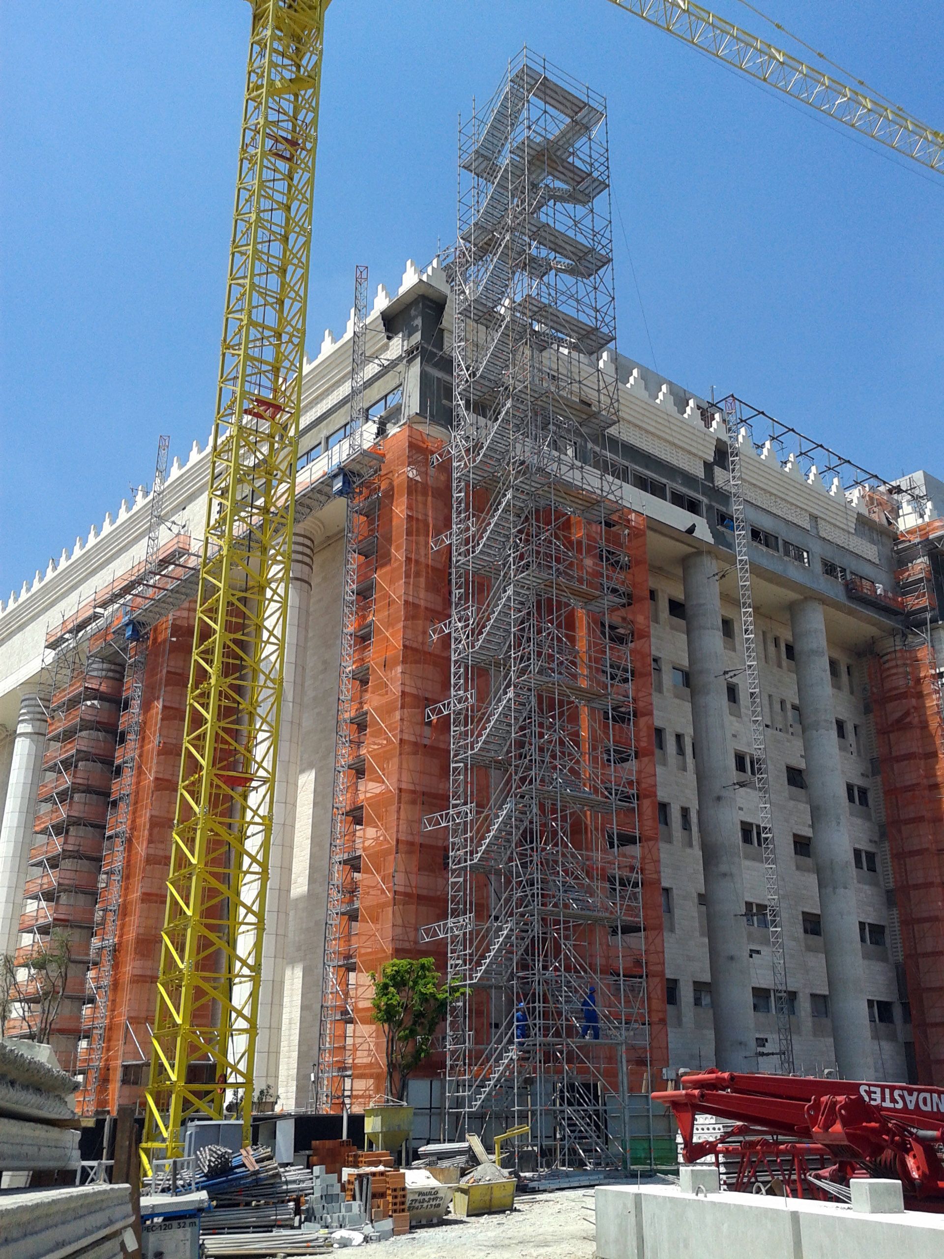 Canteiro de obras com guindastes altos e andaimes ao redor de um prédio de vários andares sob um céu azul claro.