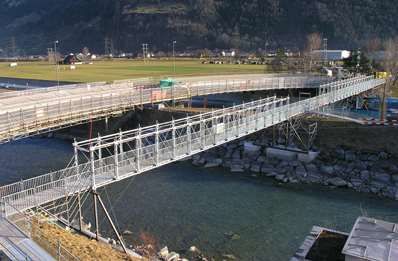Uma longa ponte pedonal branca atravessa um rio ao lado de um campo gramado e uma margem rochosa.