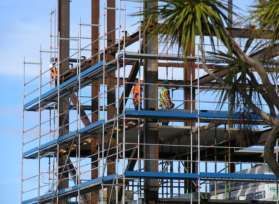 Edifício com andaimes em construção, com operários em plataformas azuis ao lado de palmeiras.