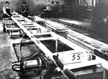 Operários montando uma longa estrutura de madeira com o número 55 em uma oficina.