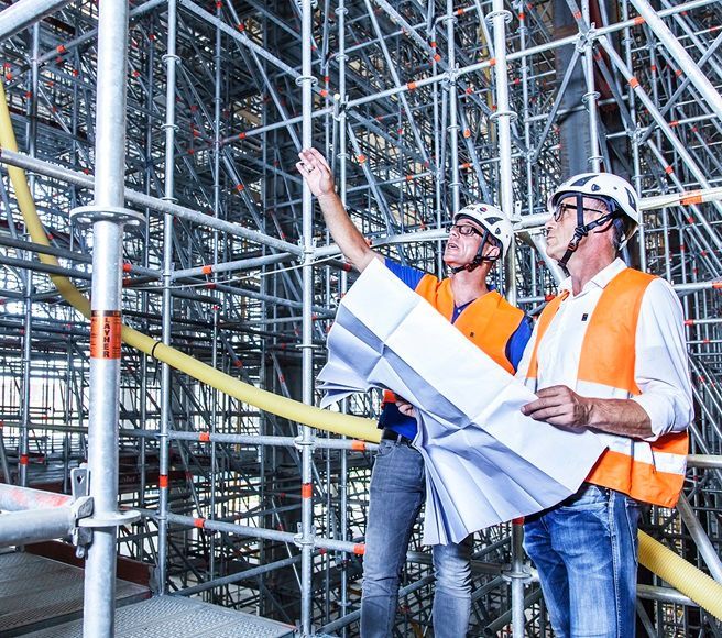 Dois operários da construção civil, usando capacetes de segurança, analisam plantas em um canteiro de obras com andaimes.