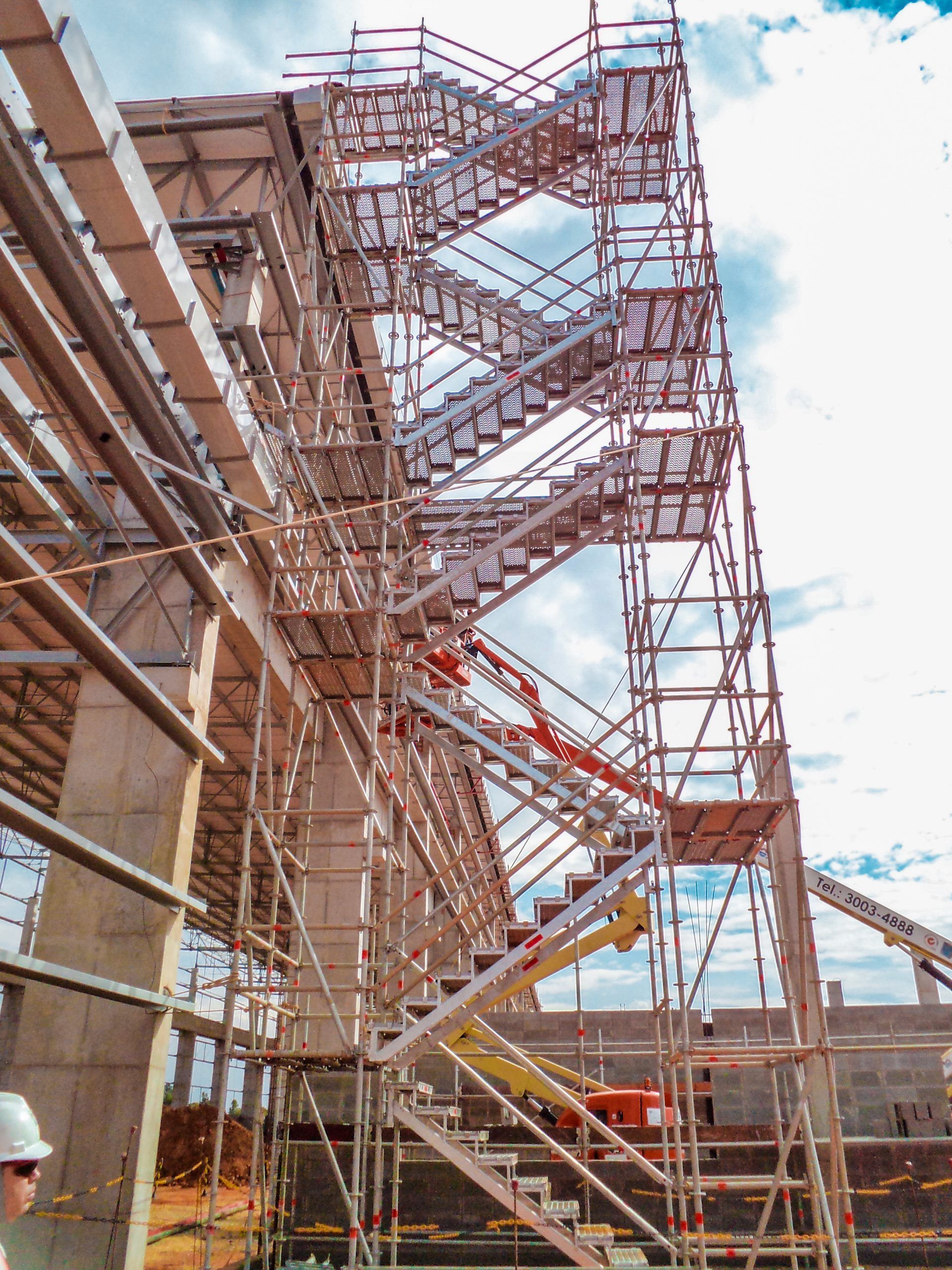 Andaimes e torre de escada metálica ao lado de um prédio de tijolos em um canteiro de obras sob um céu claro.
