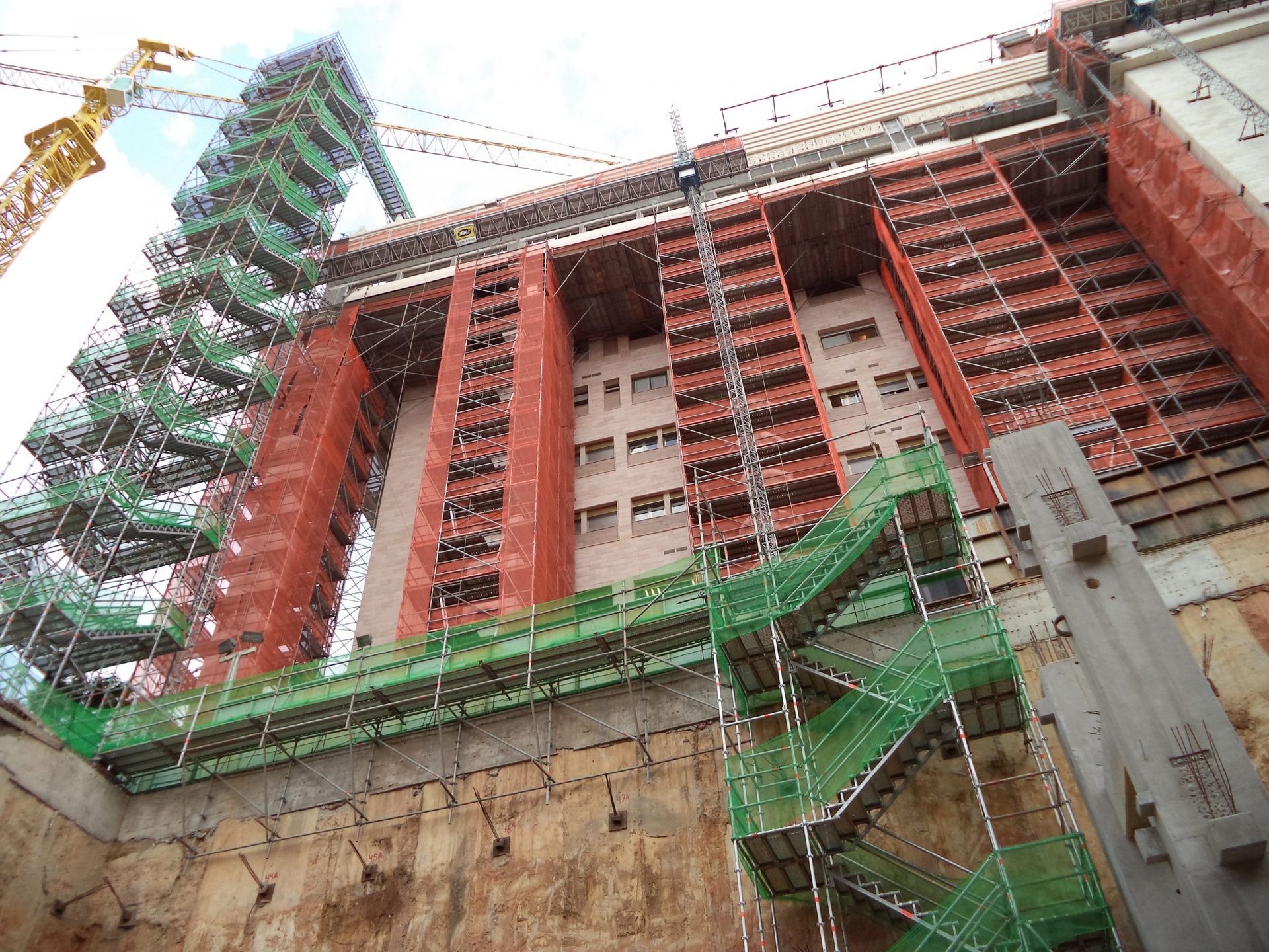 Prédio alto em construção com andaimes vermelhos e verdes contra um céu nublado.