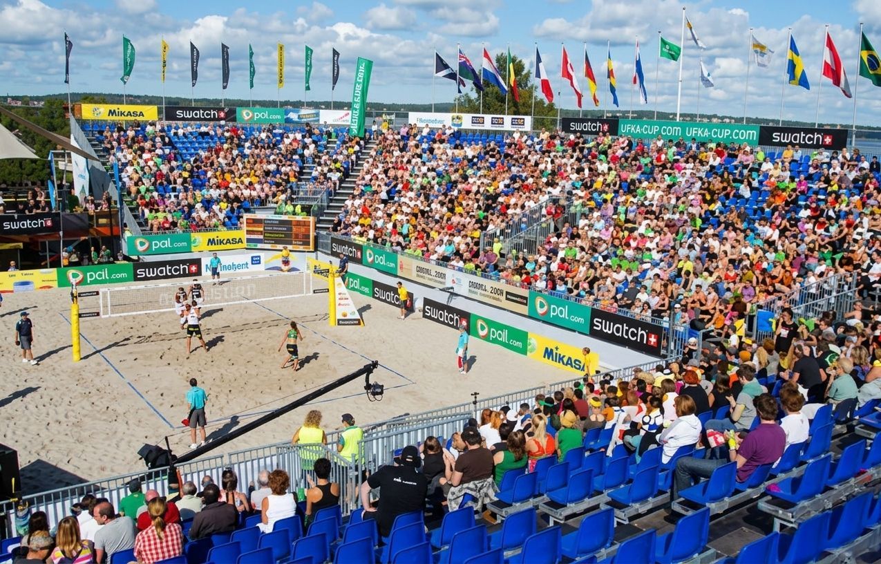 Estádio de vôlei de praia lotado, com jogadores na quadra de areia e espectadores coloridos em cadeiras azuis e laranjas.