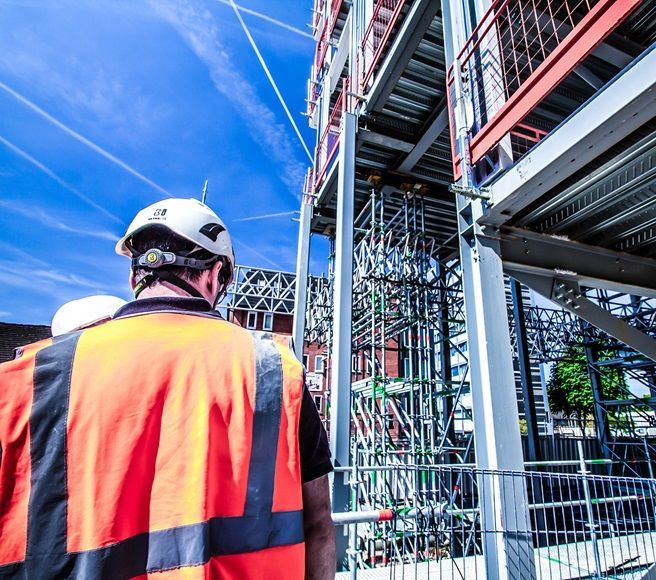 Operário da construção civil com colete laranja e capacete em uma estrutura de aço sob um céu azul brilhante.
