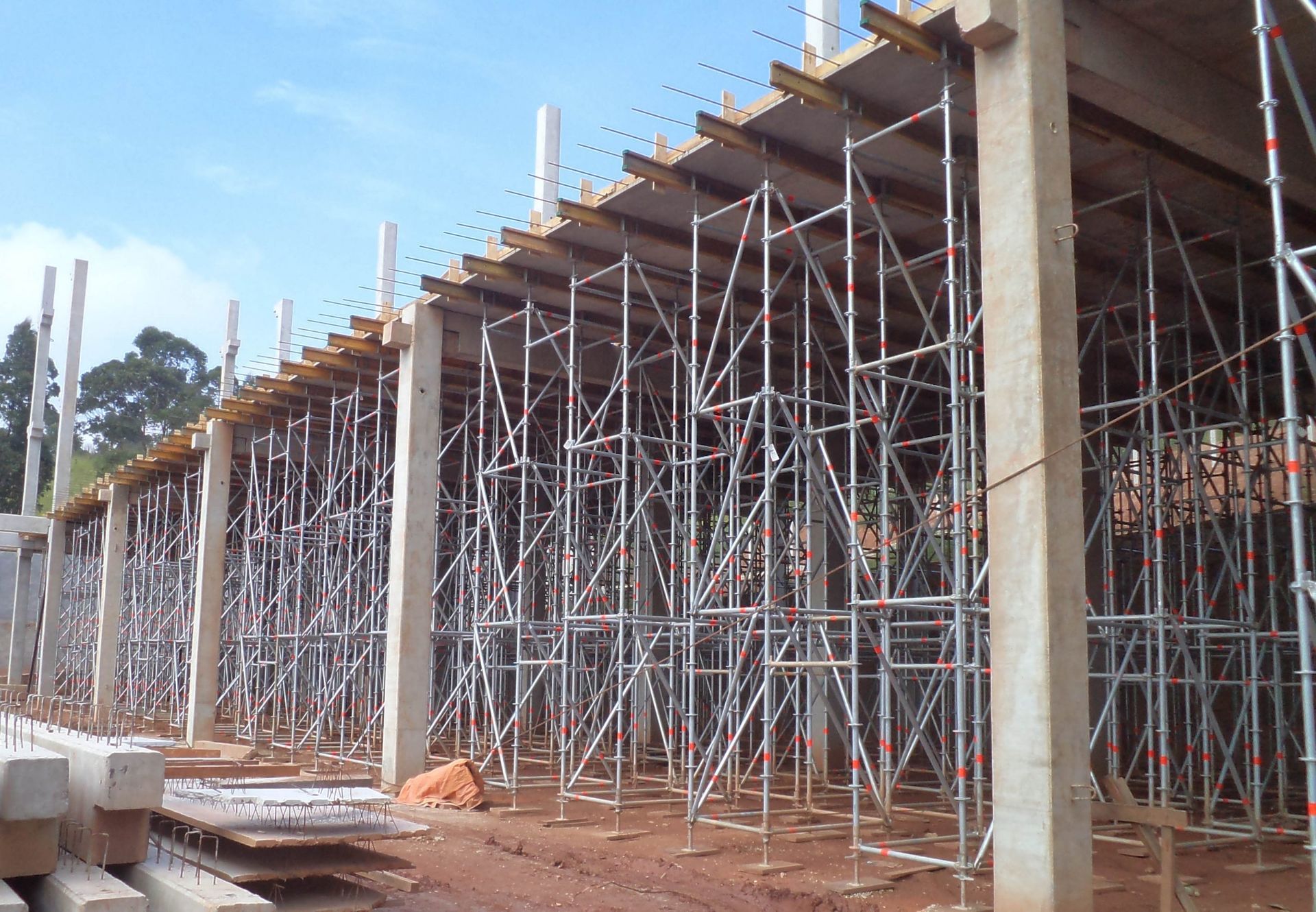 Canteiro de obras com extenso andaime metálico sustentando uma estrutura de concreto sob um céu azul.