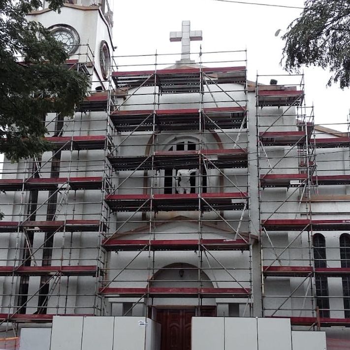 Andaimes cobrindo a fachada branca de uma igreja com uma cruz no topo e detalhes em vermelho durante a reforma.