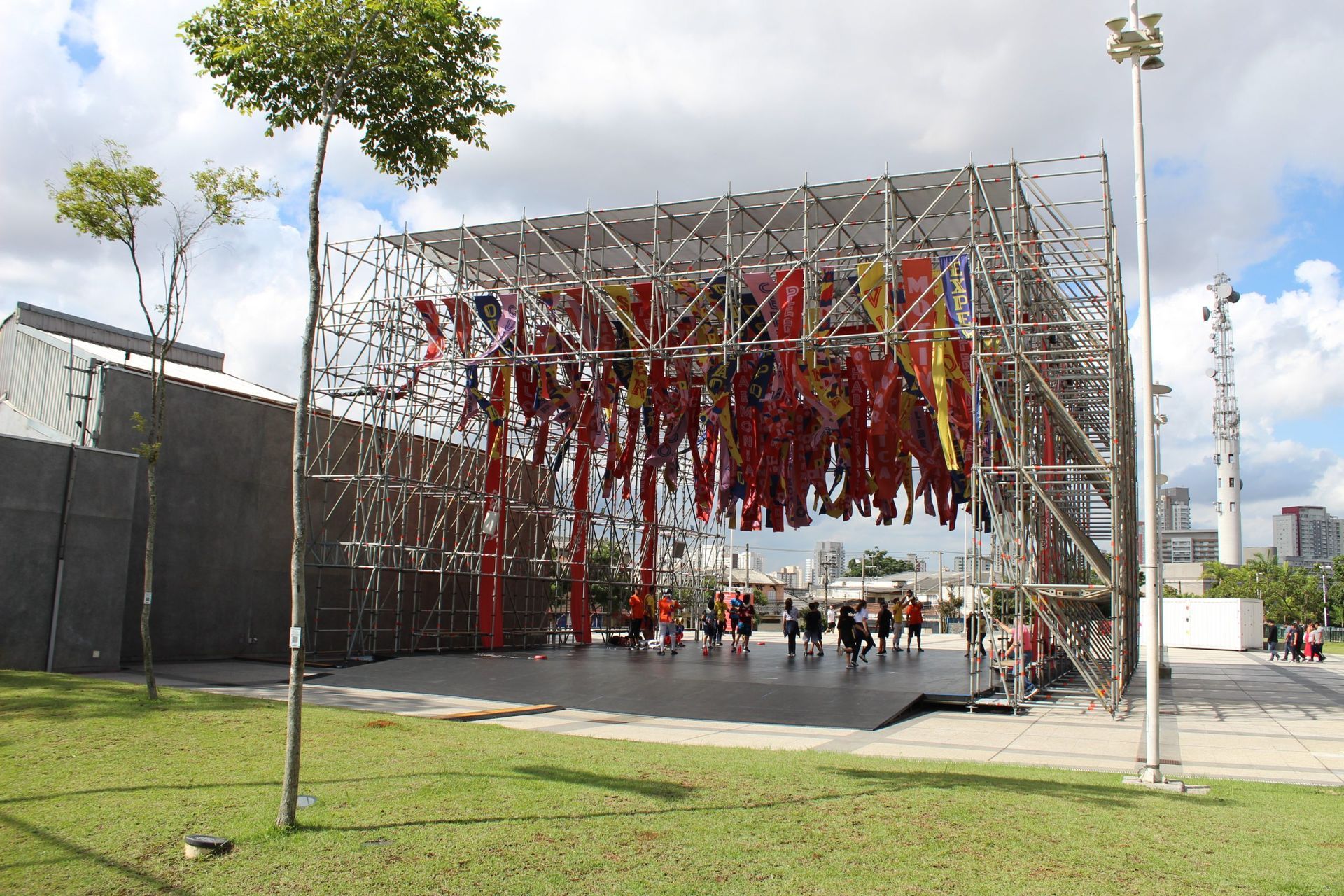 Instalação de arte ao ar livre com fitas coloridas penduradas em uma praça ensolarada perto de árvores e um prédio moderno.