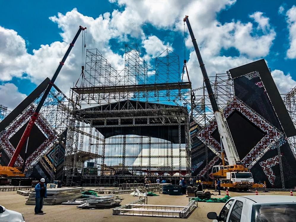 Palco de concertos em construção, com guindastes, andaimes e grandes painéis de LED pretos contra um céu azul.