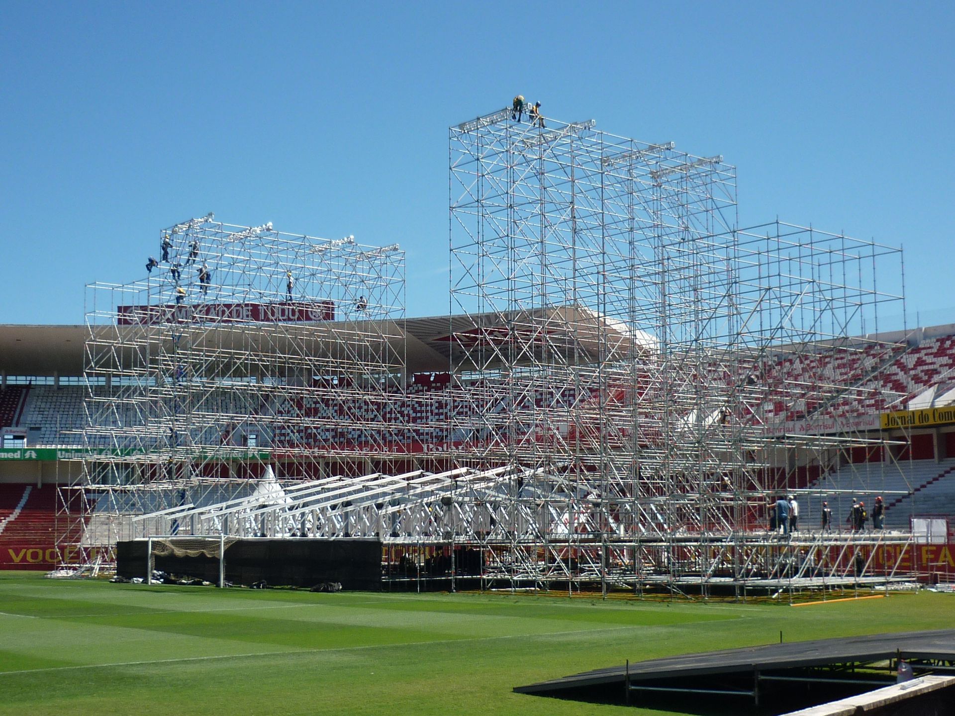 Estrutura de aço em construção em um estádio com campo verde e arquibancadas vermelhas sob um céu azul.