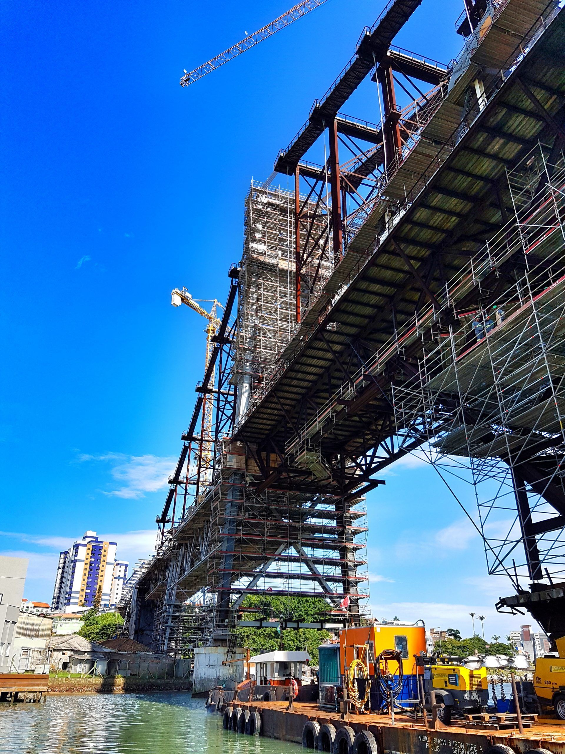 Ponte suspensa em construção sobre um rio, com andaimes, guindastes e operários abaixo.