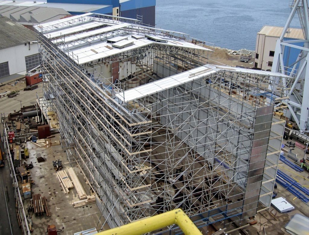 Grande estrutura industrial de aço em construção junto à água, com andaimes e guindastes.