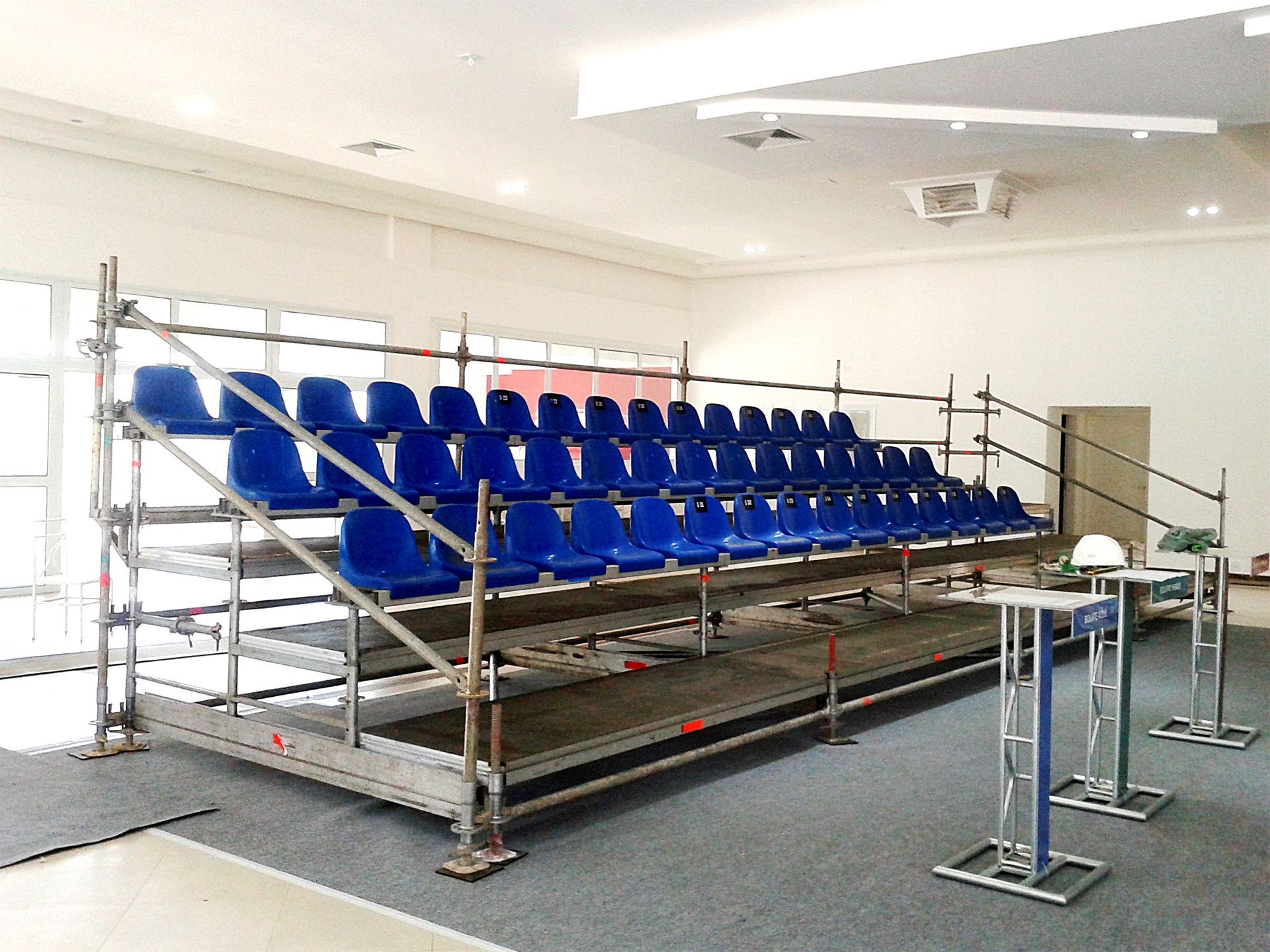 Assentos azuis estilo estádio em andaimes metálicos em uma sala interna, como arquibancadas temporárias.