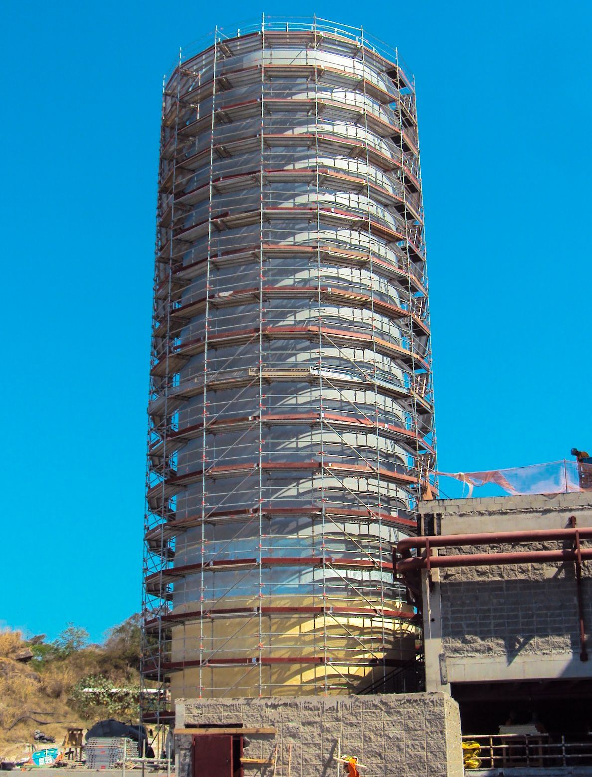 Edifício alto e cilíndrico em construção com andaimes contra um céu azul claro.