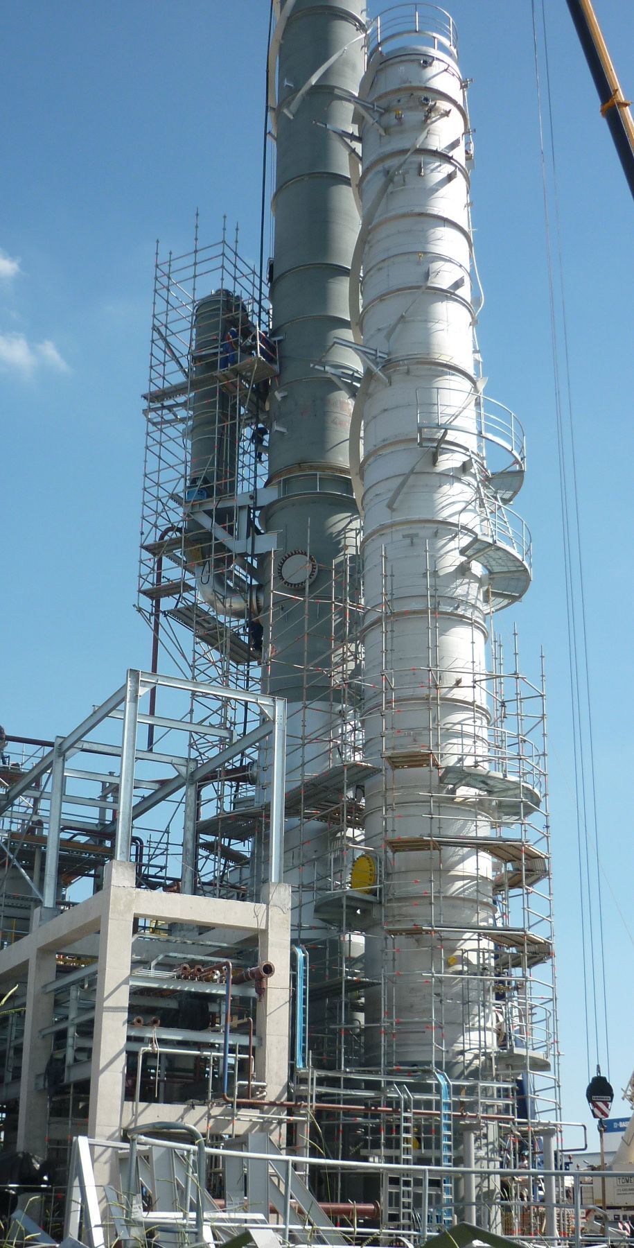 Torre de refinaria industrial com altos tubos prateados e andaimes contra um céu azul claro.