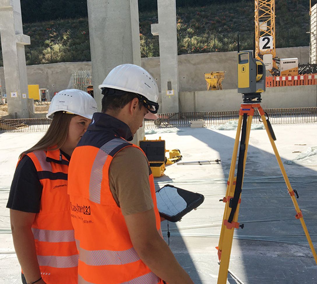 Dois operários da construção civil, com coletes laranja e capacetes brancos, utilizam equipamentos de topografia.