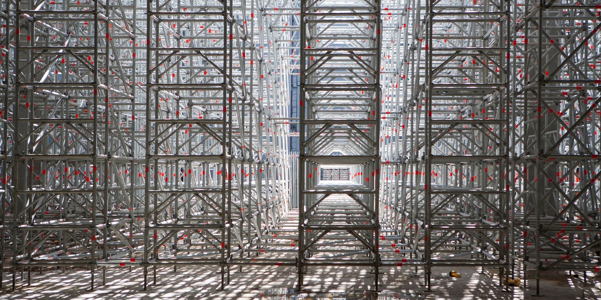 Fileiras de andaimes metálicos em um corredor de um canteiro de obras, formando um túnel industrial simétrico.