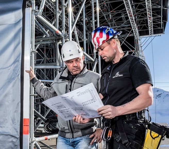 Dois operários da construção civil, usando capacetes de segurança, analisam plantas ao lado de uma estrutura de andaime.