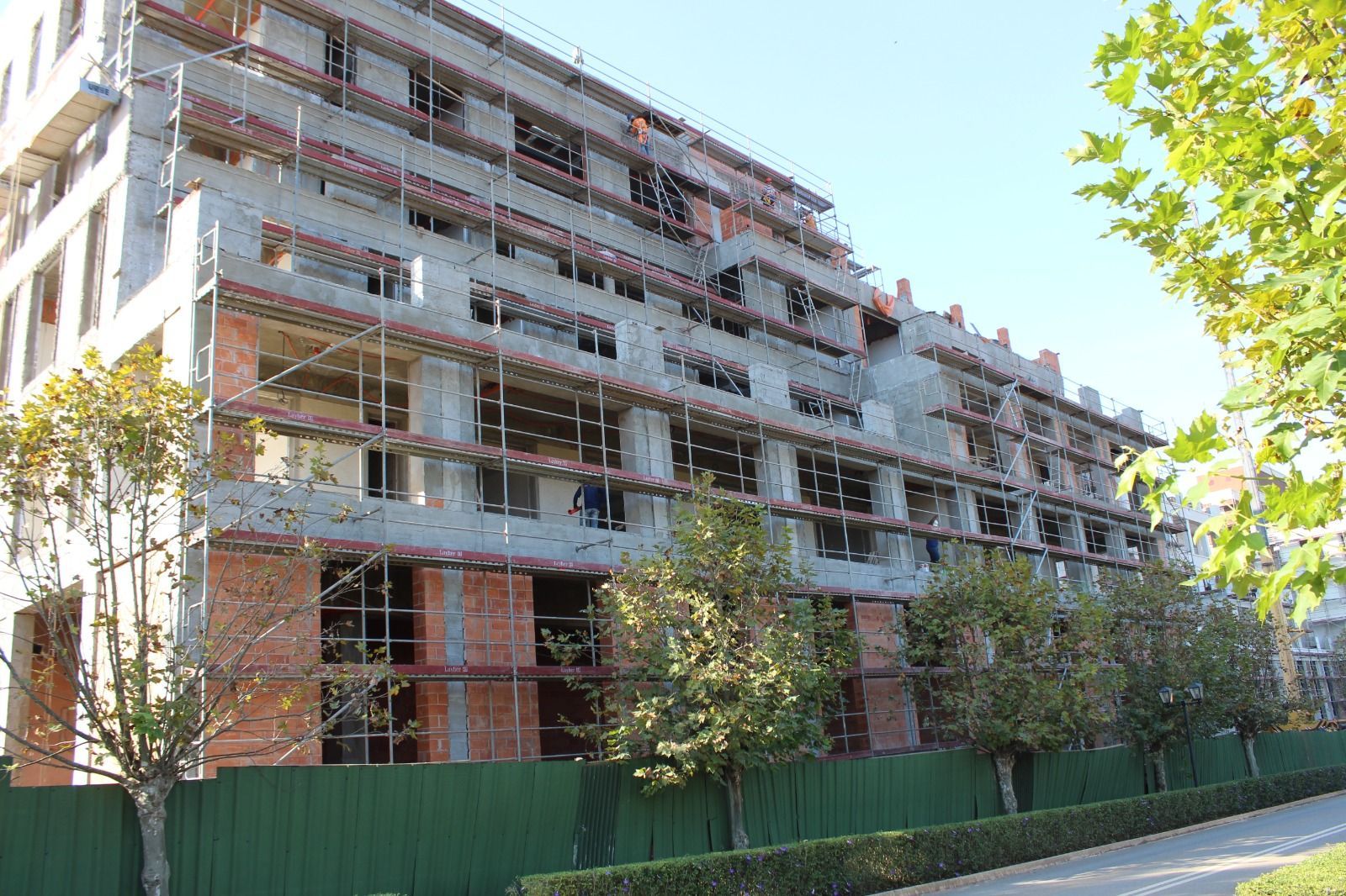 Prédio de apartamentos atrás de uma cerca verde em uma rua ensolarada, com árvores na frente.
