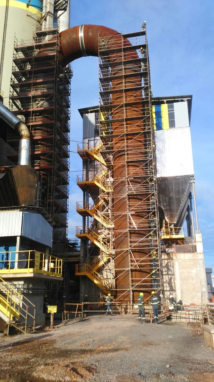 Fábrica com um grande duto curvo cor de ferrugem e andaimes ao lado de silos metálicos.
